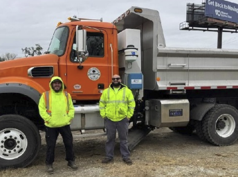 two men wearing yellow jackets standing in front of a dump truck with their hands in their pockets